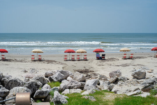 Round metal fixture and exposed pipeline emerge near large white rocks in foreground of quiet Galveston beach, adding industrial texture to scene of neatly arranged umbrellas and calm waves, TX