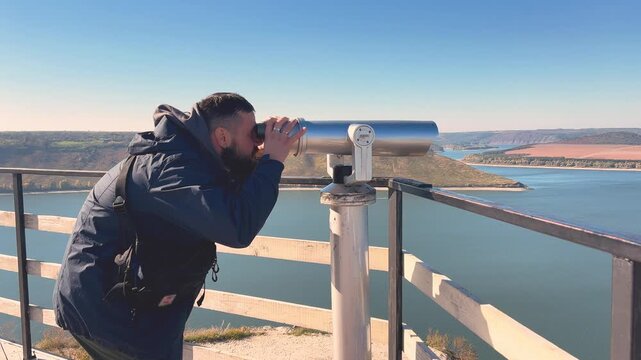Young attractive guy looking through binoculars at a mountain lake from a viewing platform