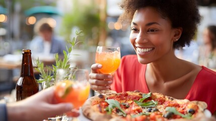 Close up of a happy woman toasting with a vibrant orange cocktail next to a delicious pizza on a table at a sunny outdoor restaurant with friends and drinks