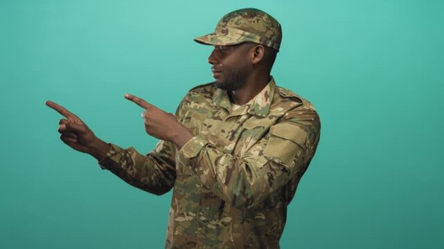Man soldier in camouflage uniform and cap points both index fingers outward while smiling in studio; confidence.