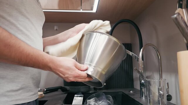 Man in gray shirt is washing a stainless steel pot in a modern kitchen sink, showcasing the cleaning process with a gradual zoom in on the action