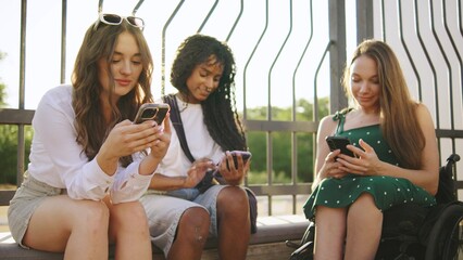 three multiracial female friends one disabled in a wheelchair engaged in digital activities outdoors during late afternoon sunlight at a park setting