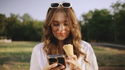 Smiling woman holds an ice cream cone and types on her phone, enjoying a warm day in a park surrounded by greenery, exuding joy and casual vibes.