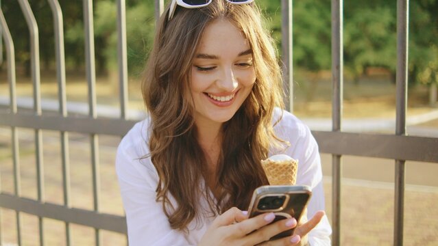 Young woman enjoys ice cream while using her smartphone in a park on a sunny afternoon, showing joy and relaxation - Powered by Adobe