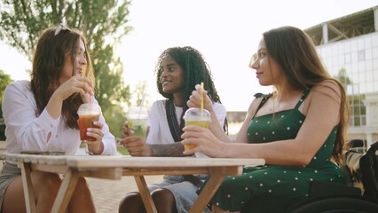 three female multiracial friends one disabled woman in a wheelchair enjoying refreshing drinks at an outdoor cafe on a sunny afternoon amidst greenery and lively conversation