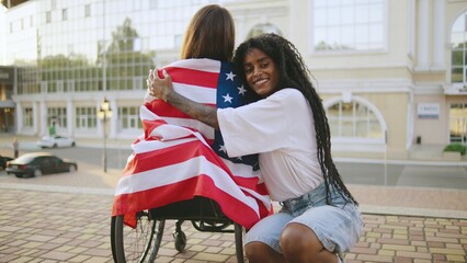 two female multiracial friends one inclusive woman in a wheelchair enjoying a sunny day in the city, celebrating freedom and friendship with an American flag