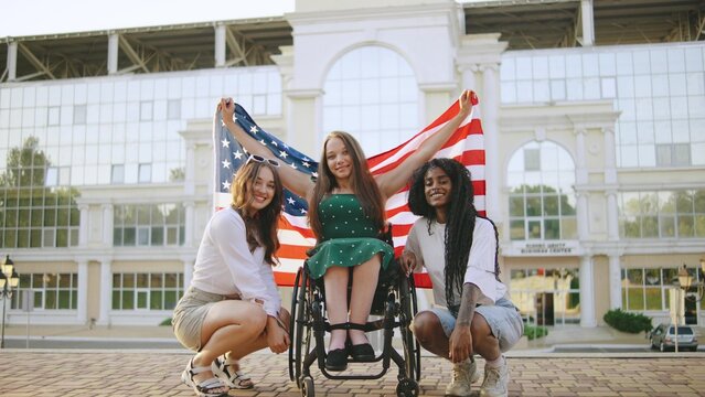 three female multiracial friends one inclusive woman in a wheelchair celebrate together while holding an American flag on a sunny day at a public park