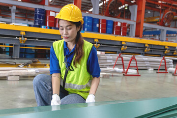 Female Asian worker with helmet and safety vest working at manufacturing factory. Woman engineer wear gloves while inspecting metal sheets in industrial site. Inspector checking the quality of product