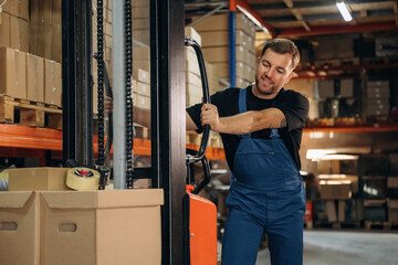 Fototapeta premium Transporting the boxes by using forklift. Man in uniform is working in the storage with packages for shipment