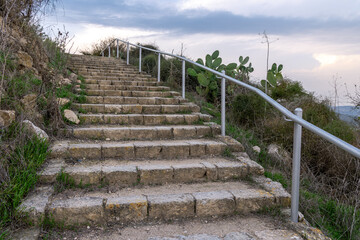 Stone stairway with metal handrail climbs a hillside path in northern Israel. Galilee park landscape with cactus, greenery and soft evening sky, inviting hiking and outdoor travel
