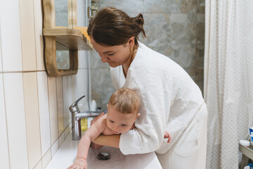 A mother helps her baby play at the sink in the bathroom. The baby reaches for the faucet while the mother supports them. Water and bath products are visible around them.