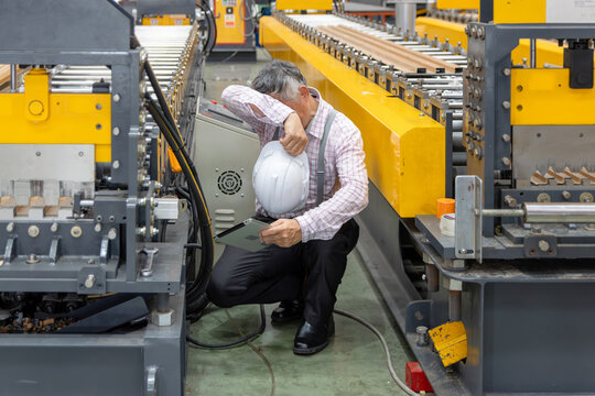 Asian senior foreman with hard hat holding tablet, wiping sweat off forehead while inspecting machinery in manufacturing factory. Tired elderly supervisor with helmet working in industrial site.