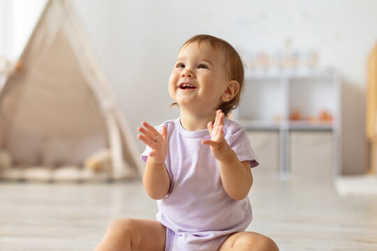 Smiling infant baby girl claps hands while sitting on floor in bright home interior. The joyful moment captures happiness, expression, and the playful curiosity of early childhood