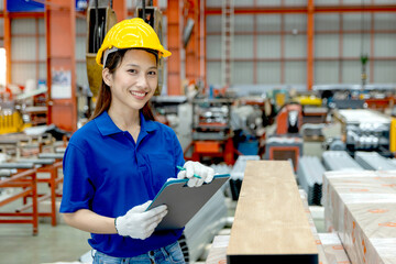 Female Asian worker with safety helmet holding a clipboard, working at manufacturing factory. Woman engineer inspecting metal sheets in industrial site. Inspector checking the quality of product.
