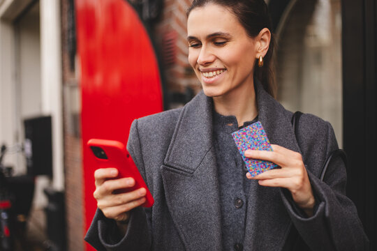 Stylish woman holding smartphone and credit card while making a mobile payment outdoors. Cashless payment, fintech, digital wallet and modern urban lifestyle concept. Happy smiling woman in grey coat.