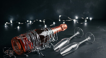 A bottle of rose liquid wrapped with silver tinsel, two glass flutes and string lights on dark textured surface, represents celebration, party