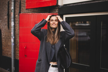 Attractive brunette woman walk on the town city street. Girl wear grey wool coat and look happy and smiles. Woman fix hair while go on the street. Girl stand near red door. 