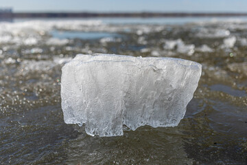 A large ice block is prominently positioned on the surface of a frozen lake, glistening under bright sunlight, surrounded by shimmering ice and water, creating a serene winter landscape