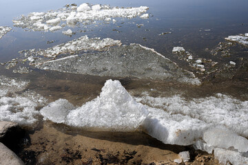 Unique ice formations are visible on the water surface, contrasting with the sandy shore. The scene captures the beauty of nature, showcasing textures and reflections in a serene environment