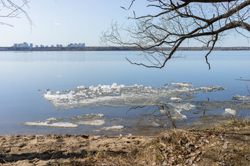 Serene lakeside scene featuring ice fragments floating on calm water, with a sandy shore and bare branches framing the view, evoking a peaceful spring atmosphere and natural beauty