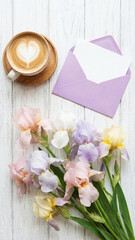 Flat lay with coffee latte art in heart foam, pastel irises, and lavender envelope with blank card on white wood. A warm, romantic scene for love notes, spring greetings, or thoughtful celebrations.