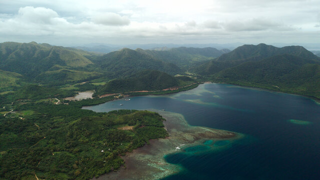 High altitude aerial panoramic view of Sitio Bayang bay and coral reef coastline near Turda in Coron, Palawan, Philippines, with lush green hills under soft overcast natural daylight.