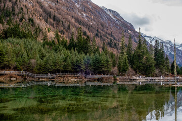 A peaceful wooden boardwalk stretches across a crystal-clear mountain lake surrounded by dense pine forests and rugged alpine slopes. The emerald-green water reflects the surrounding trees and distant