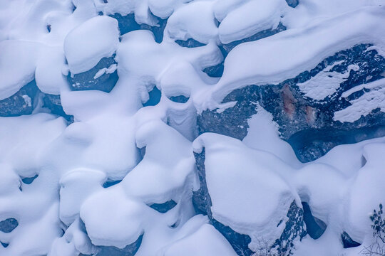 An abstract winter landscape capturing thick layers of fresh snow sculpted over rugged mountain rocks, revealing deep blue ice pools and natural organic shapes created by freezing temperatures 