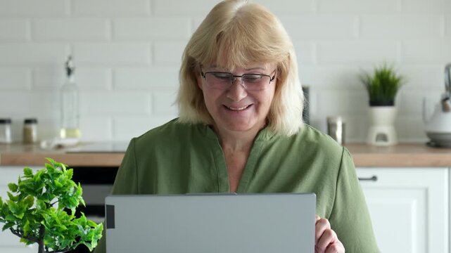 Smiling senior woman sitting in bright kitchen. Female engaging in online video call with her family or friends. Happily waving hand at webcam and communicating with loved ones