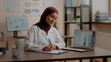 A doctor in a hijab takes notes during a virtual consultation. She is sitting at her desk, with a laptop and a clipboard Stock Video - Powered by Adobe
