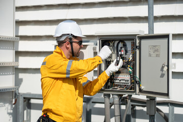 Engineer in safety helmets and yellow jackets inspecting electrical control cabinet with laptop and...