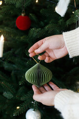 Woman decorating christmas tree with modern paper bauble, ribbons and star, hands close up. Merry Christmas and Happy Holidays! Stylish xmas tree with ornaments and lights