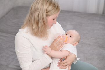 Top-down view of loving mother and baby. Mom cares for newborn baby, feeding him with bottle. Infant drinks milk while held in mother's arms. Childhood, infancy, parenting, motherhood