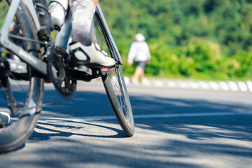 A cyclist's foot on the pedal, captured in motion on a sunny day, with a blurred background of greenery and a person walking nearby.