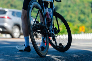 A close-up view of a bicycle wheel with a cyclist's leg in motion, set against a blurred background of a road and greenery.