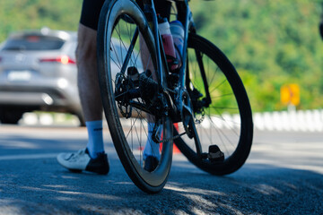 A close-up view of a bicycle wheel on the road, with a cyclist's leg visible, showcasing the surrounding landscape and a vehicle in the background.