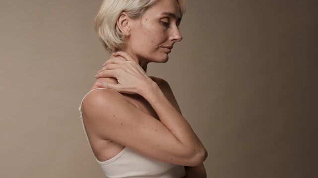 Side view of blonde mature woman in creamy sport bra turning head to camera while massaging neck muscles slowly