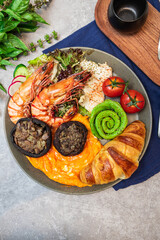 A vibrant brunch plate with shrimp, greens, avocado, mushrooms, scrambled eggs, mashed potatoes, and croissant.