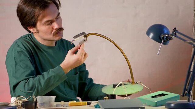 Thoughtful man restorer in creative renewal process of old item, holding inspecting vintage lamp base without lampshade, examines and evaluates condition of base parts, wires and light bulb holders.