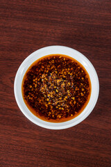 A rich red chili oil with flakes and seeds, served in a white bowl on a dark wood surface, perfect for adding heat and flavor to dishes.