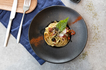 Linguine pasta in a black bowl garnished with cherry tomatoes, olives, capers, basil, radish, and red seasoning powder.