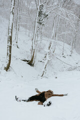 A playful, top-down shot of an unrecognizable person dressed in a warm brown coat and black pants, lying flat on their back in deep, untouched white snow. The pose is typical of making a snow angel.