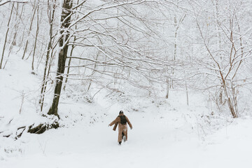 A wide shot of a solitary figure, viewed from behind, wearing a long brown coat and a black backpack, walking away from the camera into a pristine, deep, snow-covered forest clearing.