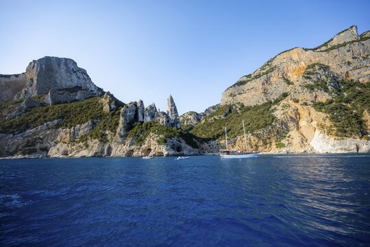 Excursion boat off picturesque rocky coast, cliffs with caves in morning light, L'Aguglia rock pin, blue sea and Cala Goloritz&eacute; beach, Golfo di Orosei, Baunei, Sardinia, Italy