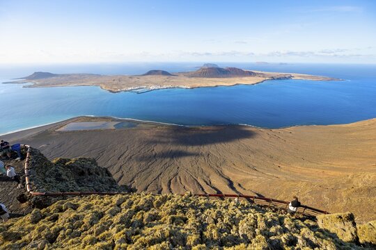 View of La Graciosa island with volcanic craters in the evening light, tourists on an observation deck at the Mirador del R&iacute;o viewpoint designed by artist C&eacute;sar Manrique, Lanzarote, Canary Islands, Spain