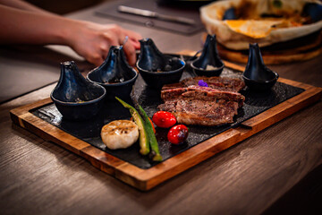 A gourmet grilled steak served on a rustic black slate board with assorted sauces, roasted garlic, cherry tomatoes, and okra. The rich texture and elegant plating highlight a premium dining experience