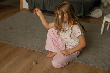 Little Girl Playing With Spinning Top On Hardwood Floor In Pajamas, Focused Expression, Pulling String To Launch Toy, Vintage Wooden Top, Soft Morning