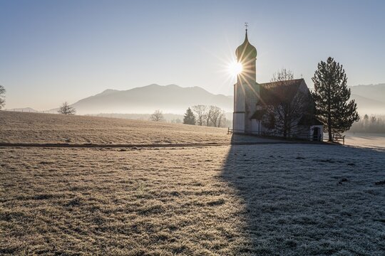 Church backlit with sun in front of mountains, winter, fog, hoarfrost, Sankt Johannisrain, Penzberg, Alpine foothills, Zwiesel in the background, Bavaria, Germany