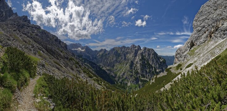 A narrow mountain path winds its way through a landscape of lush greenery and rock faces under a cloudy sky, Zugspitze, Waxensteinspitze, Höllental, Hammersbach, Osterfelderkopf, Kreuzeck, Garmisch-Partenkirchen, Wetterstein range, Bavaria, Germany - Powered by Adobe