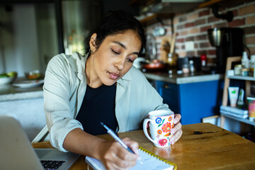 Young adult woman focused, writing notes at home kitchen table